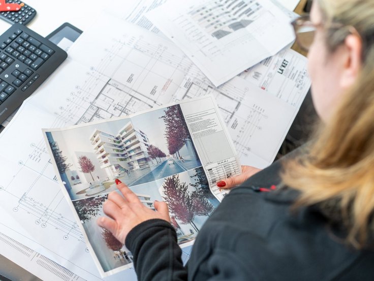 Frau zeigt auf ein Foto eines Neubauprojektes- im Hintergrund liegen Grundrisse und Zeichnungen dieser Immobilie