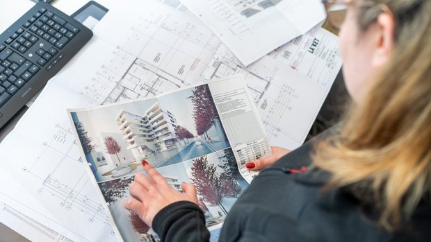 Frau zeigt auf ein Foto eines Neubauprojektes- im Hintergrund liegen Grundrisse und Zeichnungen dieser Immobilie